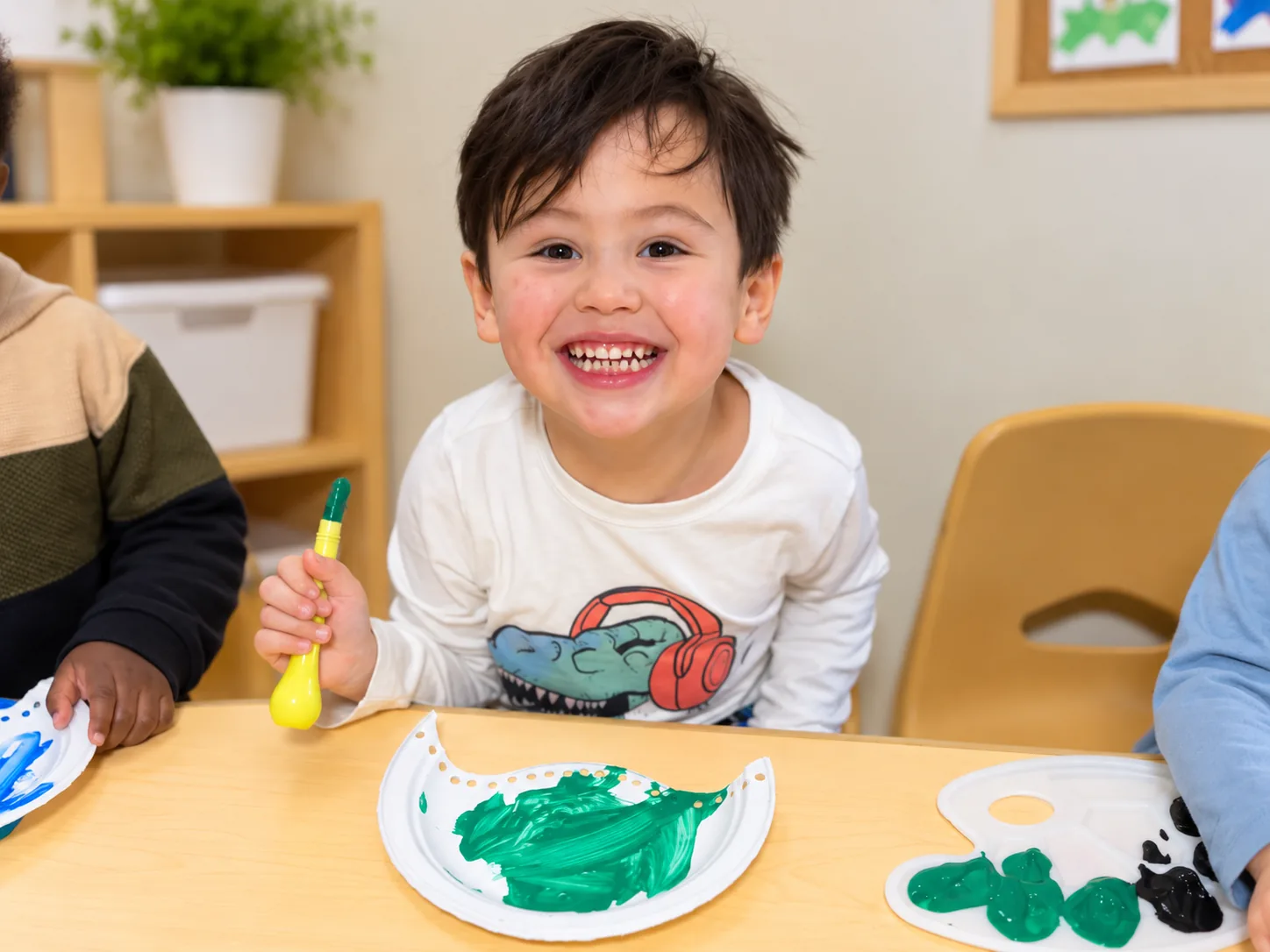 Child smiling while painting a paper plate craft with green paint at the table