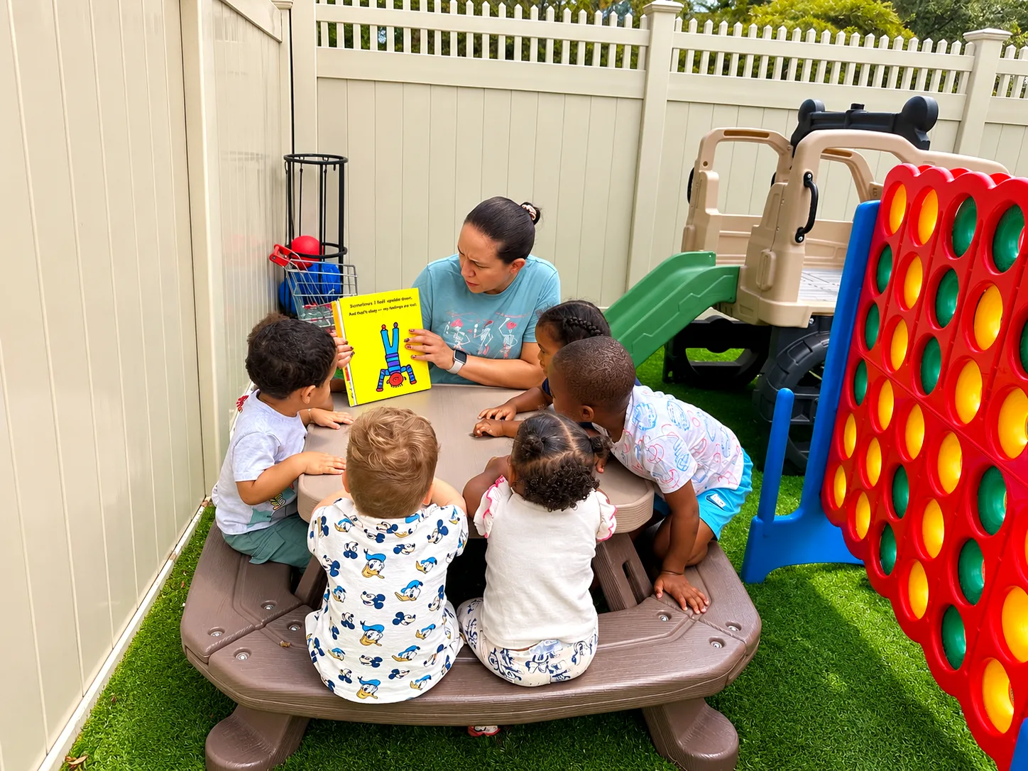 Teacher reading a picture book to a group of children gathered around an outdoor table