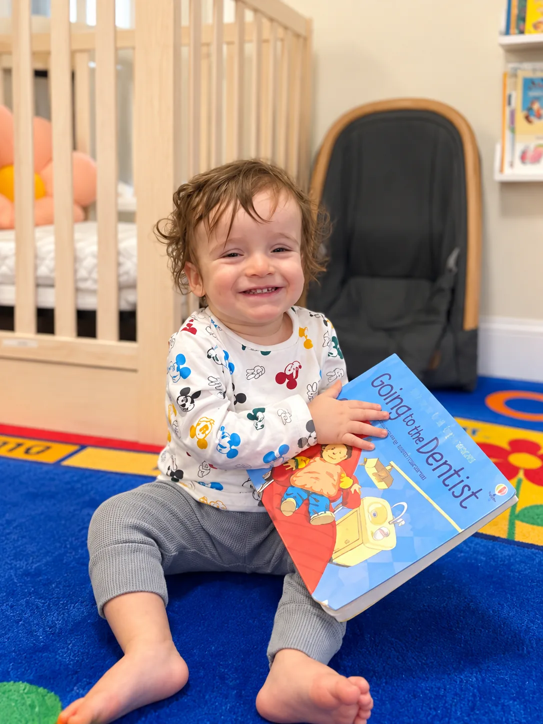 Smiling toddler sitting on a play rug holding a picture book in the infant room
