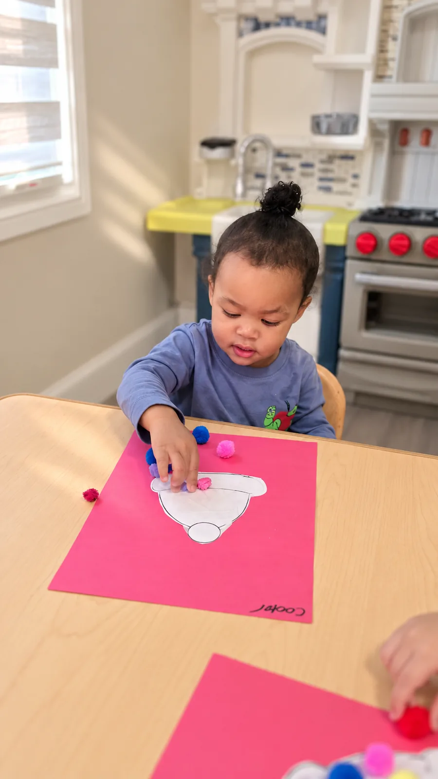 Toddler placing colorful pom-poms on a paper craft over bright pink cardstock