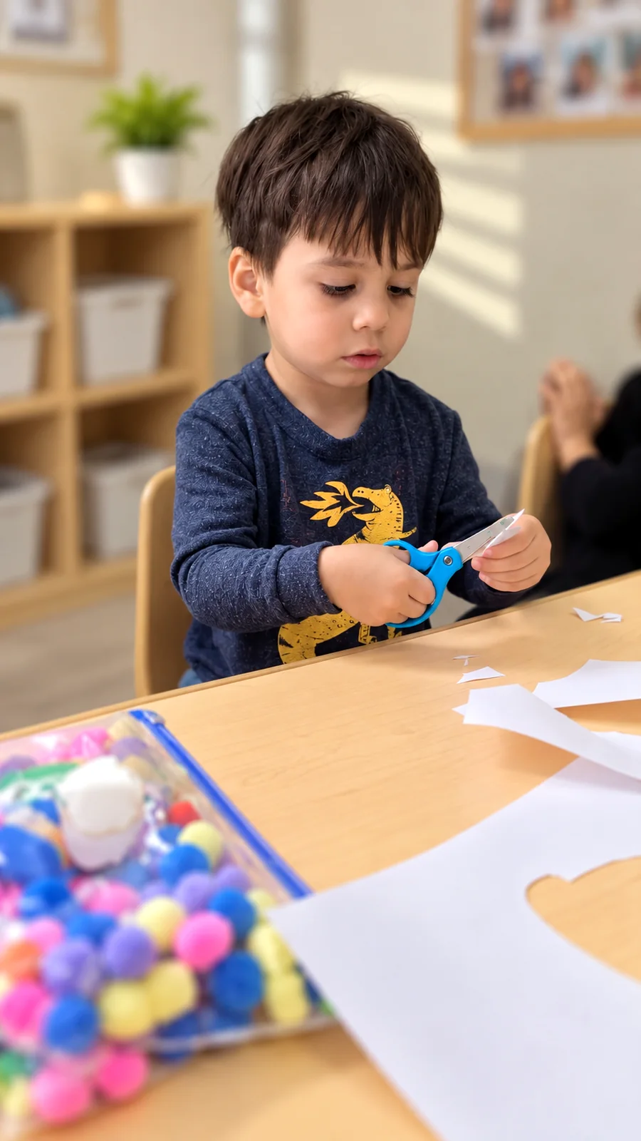 Child carefully using safety scissors beside colorful pom-poms and art materials