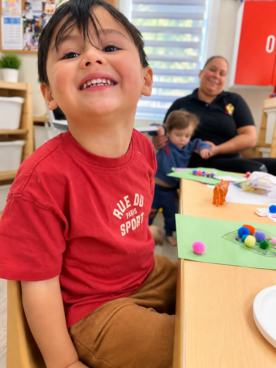 Happy child at the craft table with pom-pom projects and a teacher nearby