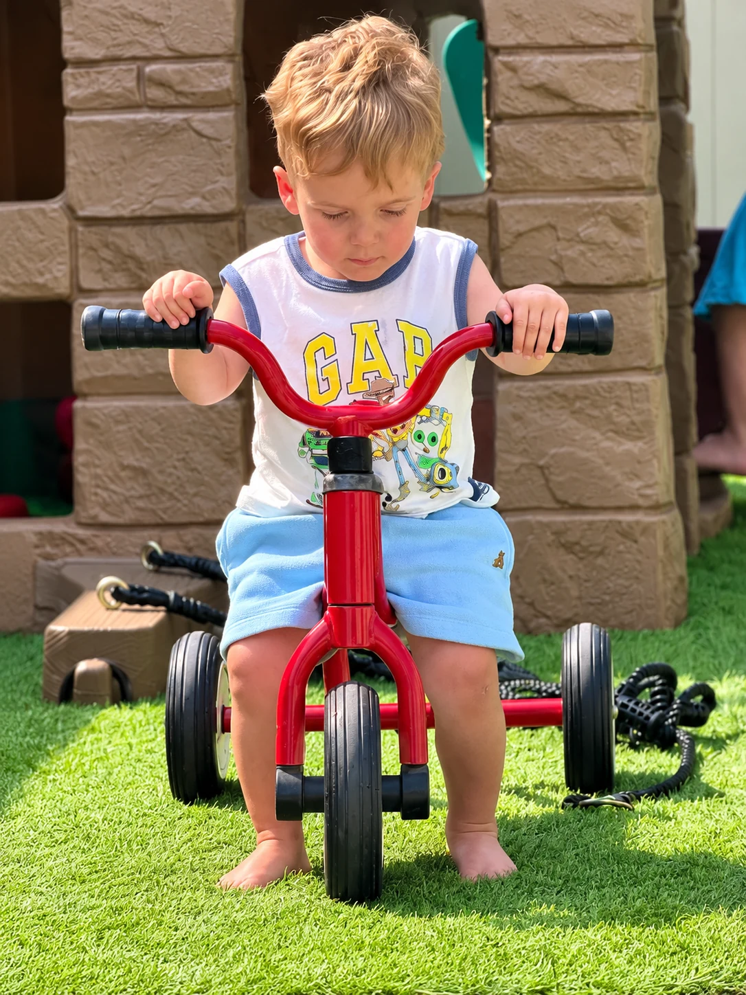 Child riding a red tricycle across the soft artificial grass play yard